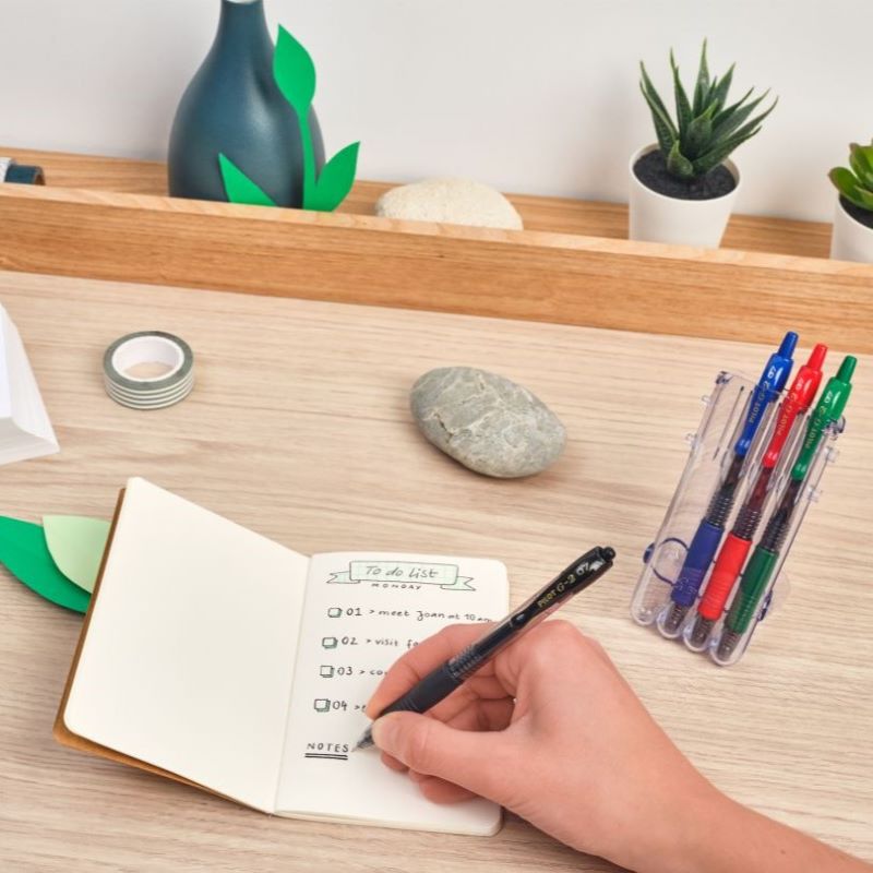 Person Writing on a Book with a Pilot G-2 Gel Pen, While Other Pens are Arranged on a Stand on the Side.